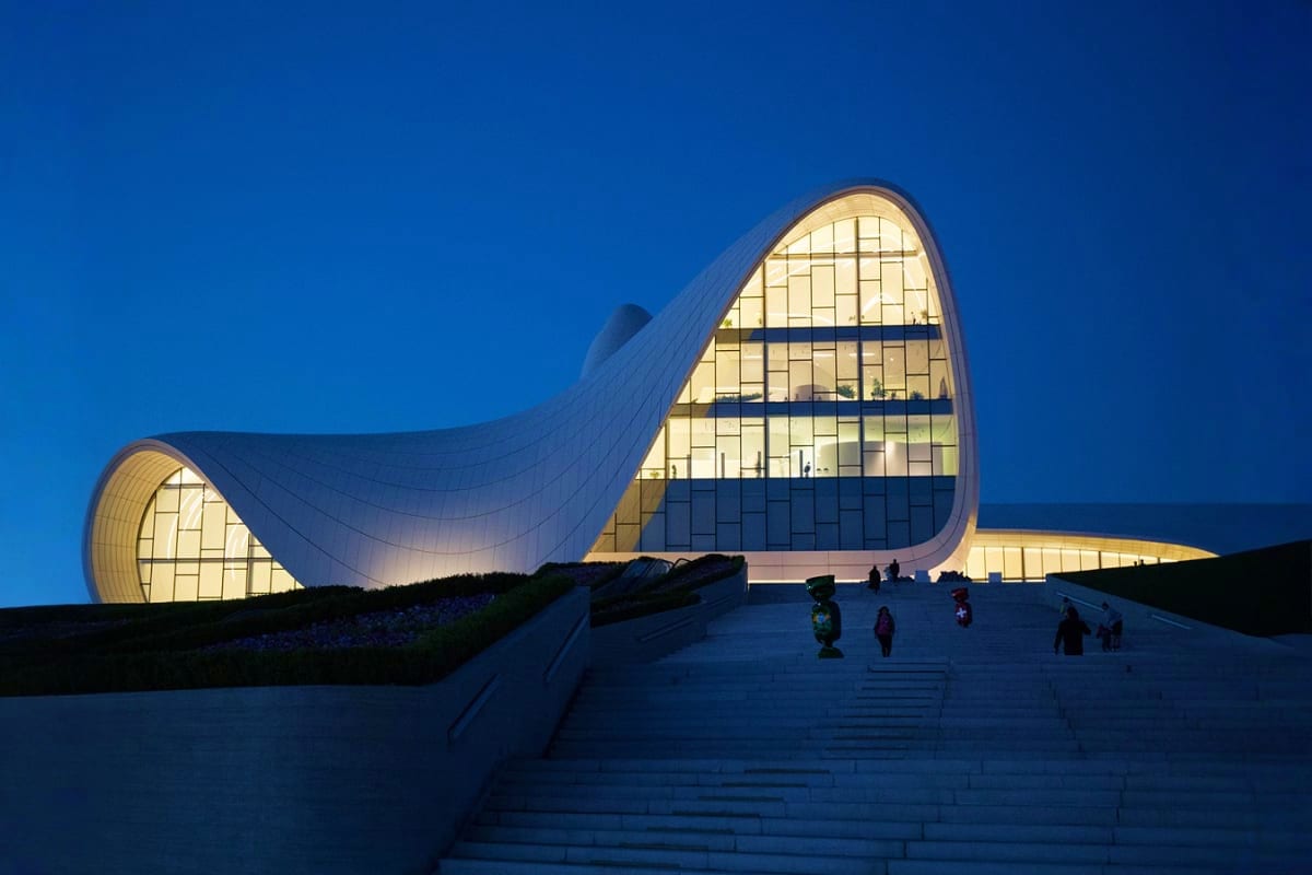 Modern illuminated building with curved white architecture and large arched windows at dusk, visitors on plaza stairs at nigh