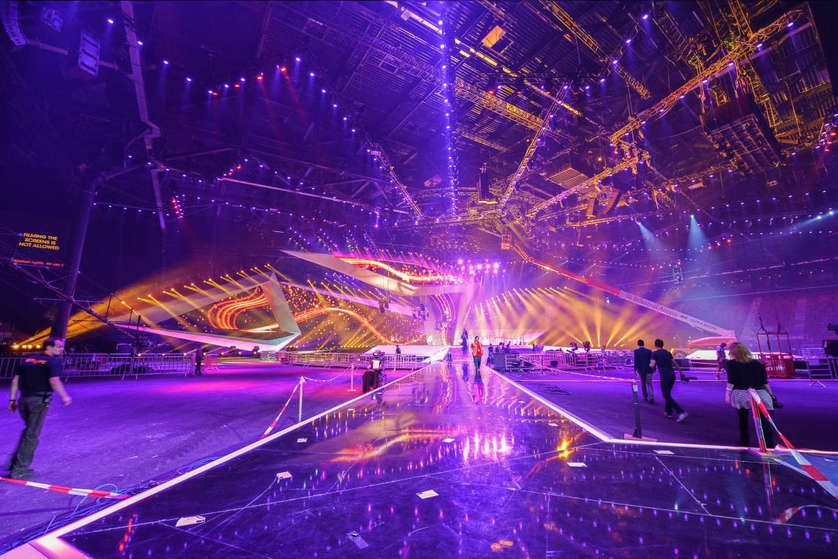 Illuminated indoor arena at night with purple and gold LED lights, reflective floor, stage lighting, and visitors walking thr