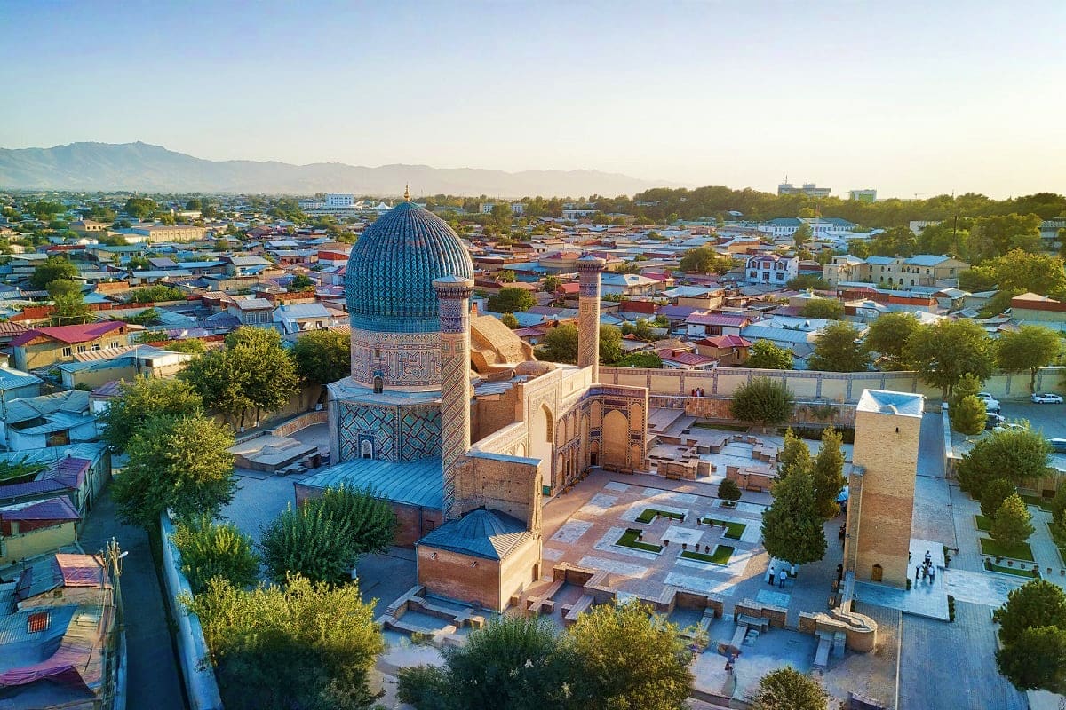 Aerial view of Imam Bukhari Complex with blue dome and minaret surrounded by Samarkand cityscape and mountains at sunrise