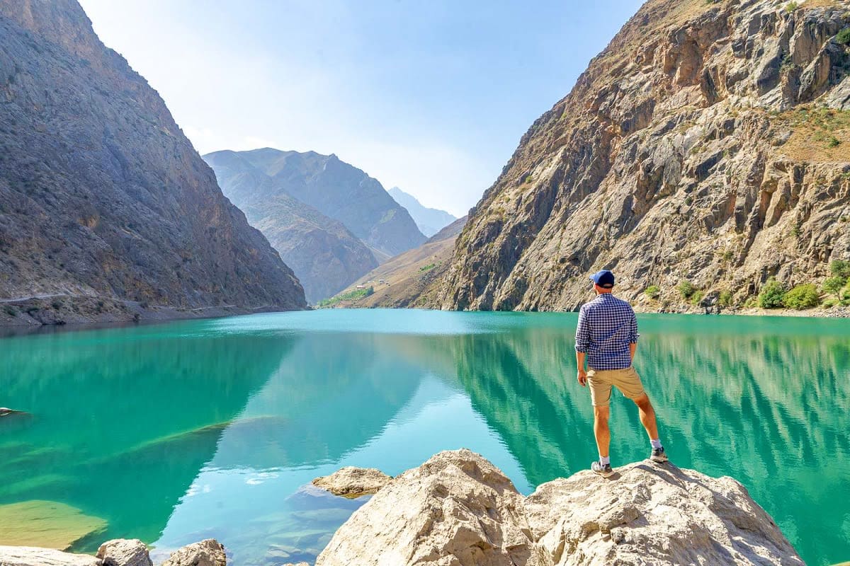 Hiker standing on rocks overlooking turquoise alpine lake surrounded by steep mountain cliffs in Tajikistan