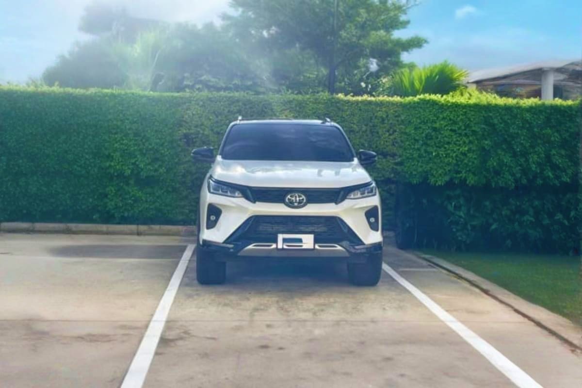 White Toyota SUV parked on driveway with green hedge fence and residential buildings in background