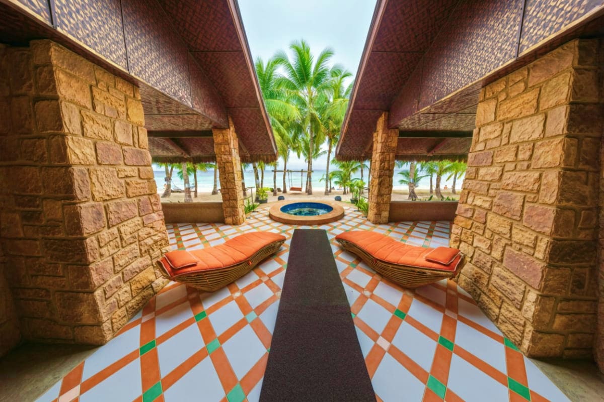 Luxury beachfront resort courtyard with symmetrical stone columns, orange loungers, central fire pit, and palm trees overlook