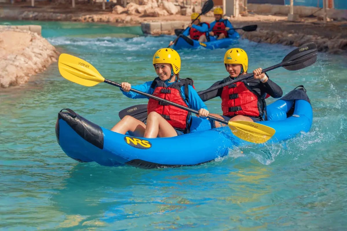 Two children kayaking in a blue NRS tandem kayak in turquoise water wearing yellow helmets and red life jackets near Abu Dhab