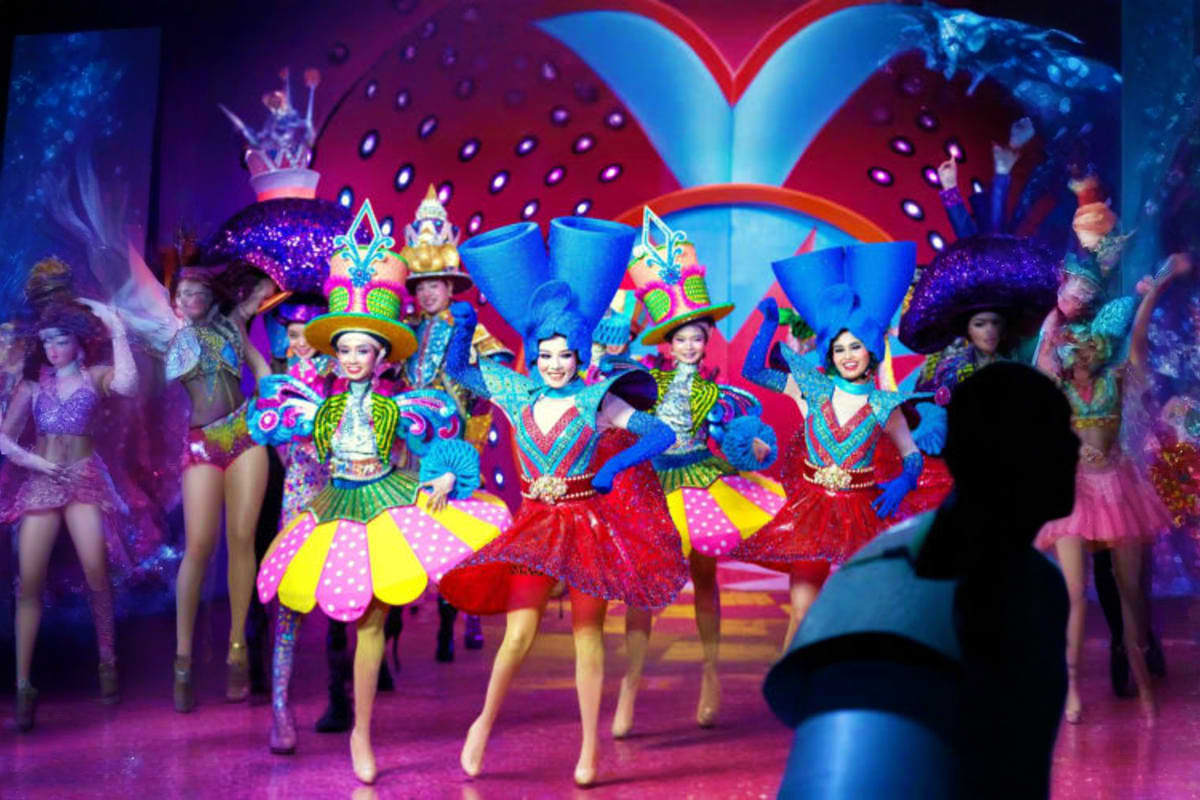 Colorful stage performance with performers in vibrant costumes and oversized headdresses dancing on a purple-lit stage with l