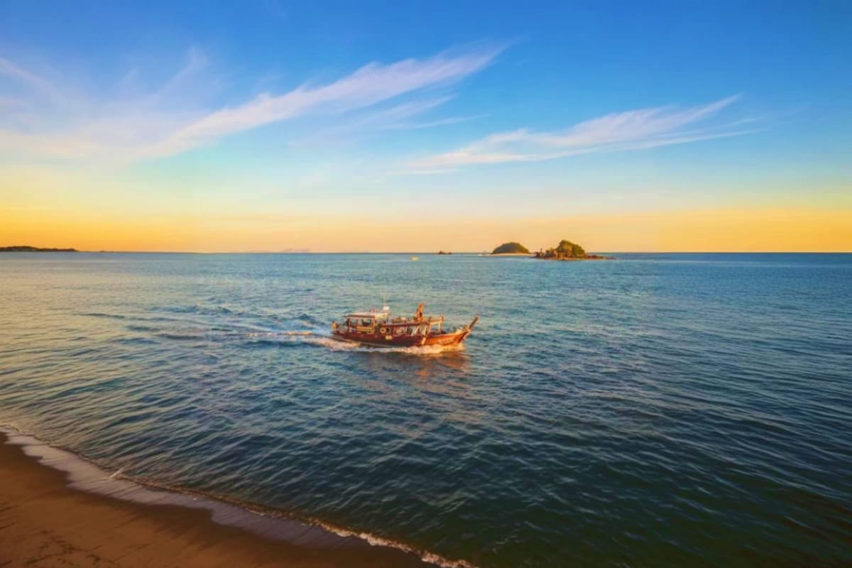 Traditional wooden longtail boat anchored in shallow water at sunset with distant islands and beach foreground