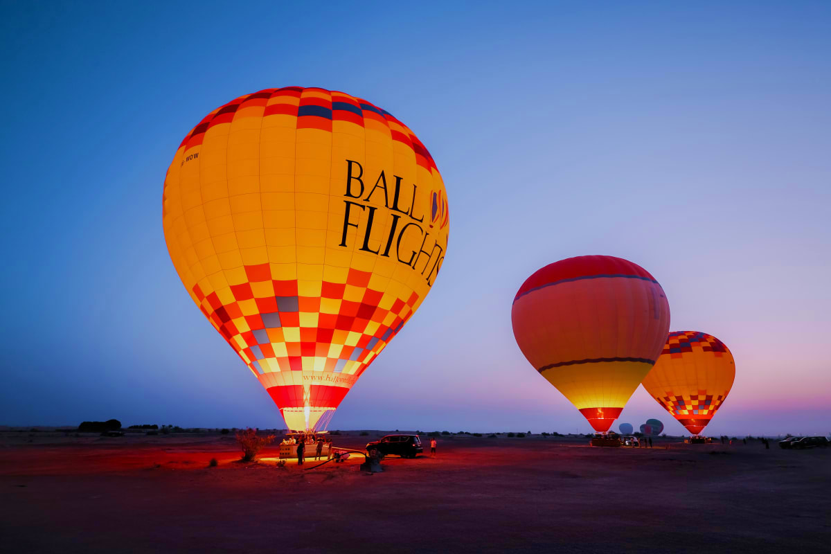Hot air balloons illuminated at dawn on flat desert terrain with ground crew and vintage vehicle nearby