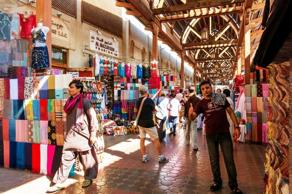 Covered marketplace in Dubai with colorful textile stalls, wooden lattice ceiling, and shoppers browsing fabrics and clothing