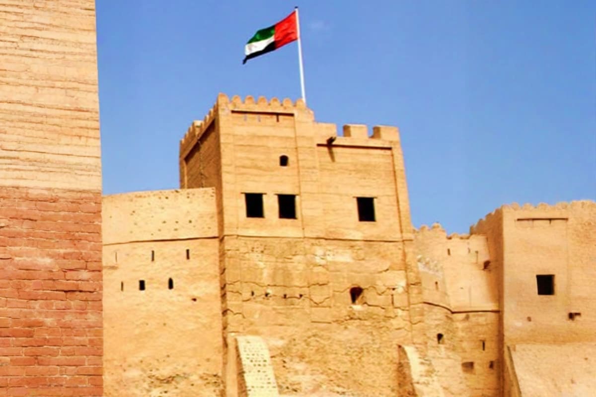 UAE flag flying above historic clay-built fort tower with crenellated walls and small windows against blue sky