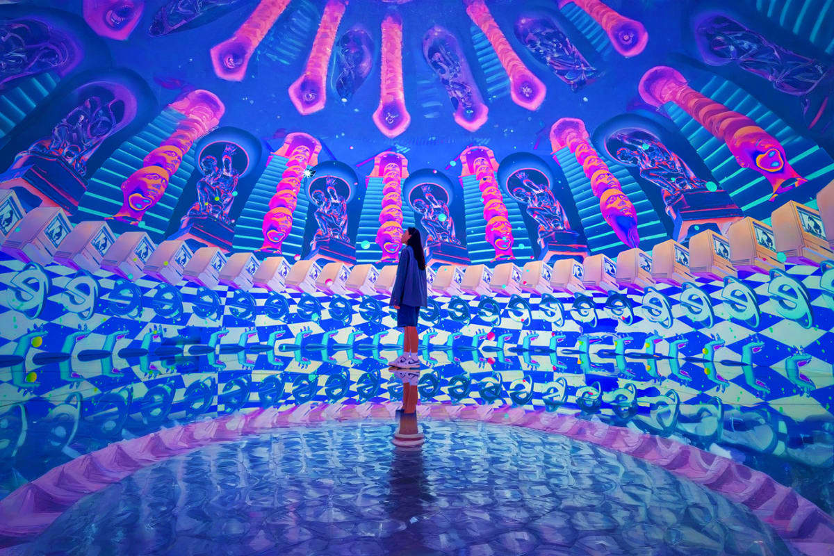 Person standing in immersive neon art installation with glowing pink and blue jellyfish-shaped sculptures overhead and reflec