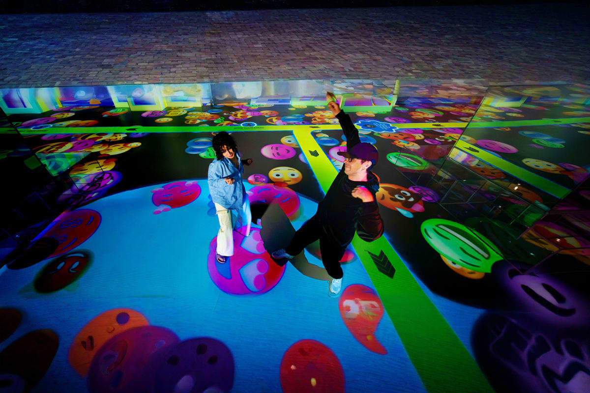 Two visitors interacting in an immersive digital art installation with colorful projected patterns and abstract designs cover