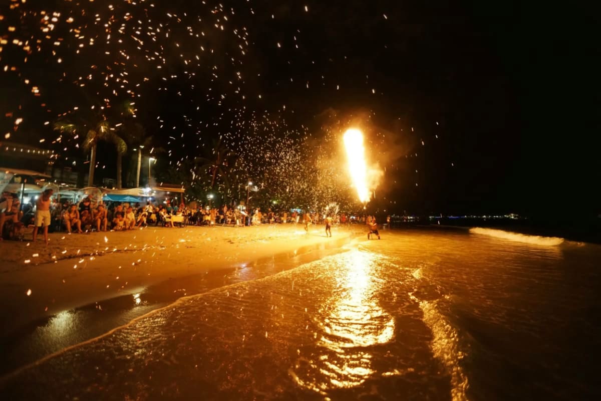 Fire breather performing at night beach party with crowds of people gathered under sparks on Koh Phangan sand