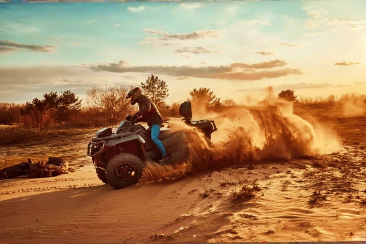 Person riding ATV through desert sand creating dust clouds at sunset with sparse vegetation and trees in background