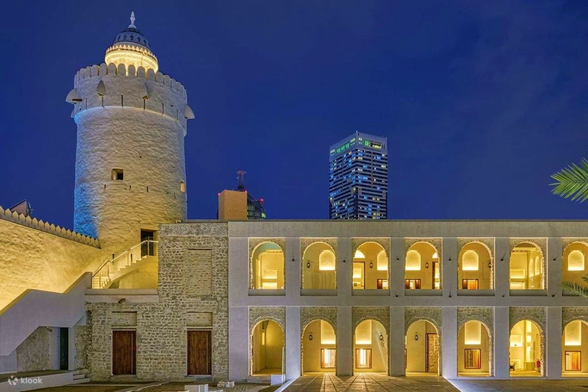 Illuminated Al Hosn fortress complex at dusk with historic minaret tower, modern courtyard with arched colonnades, and contem