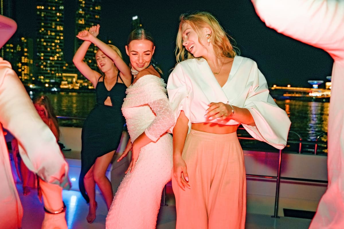 Women dancing and celebrating at a yacht party on Dubai Marina at night with illuminated city skyscrapers reflected in the wa