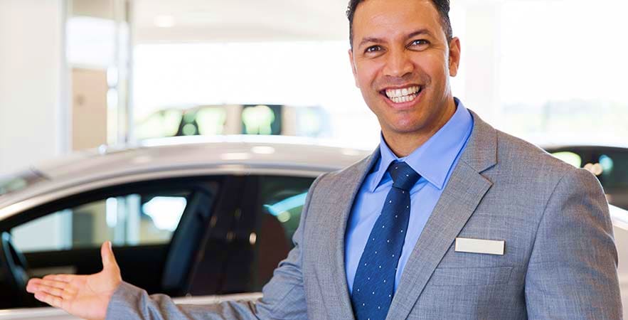 Smiling car salesman in gray suit and blue shirt standing next to vehicle in dealership showroom