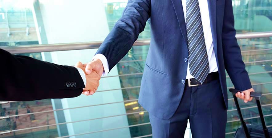 Two businessmen shaking hands in a high-rise office with city skyline visible through glass windows behind them