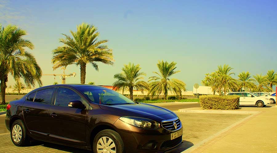 Abu Dhabi to Dubai Intercity transfers Black sedan parked in desert landscape with palm trees and modern buildings under clear blue sky