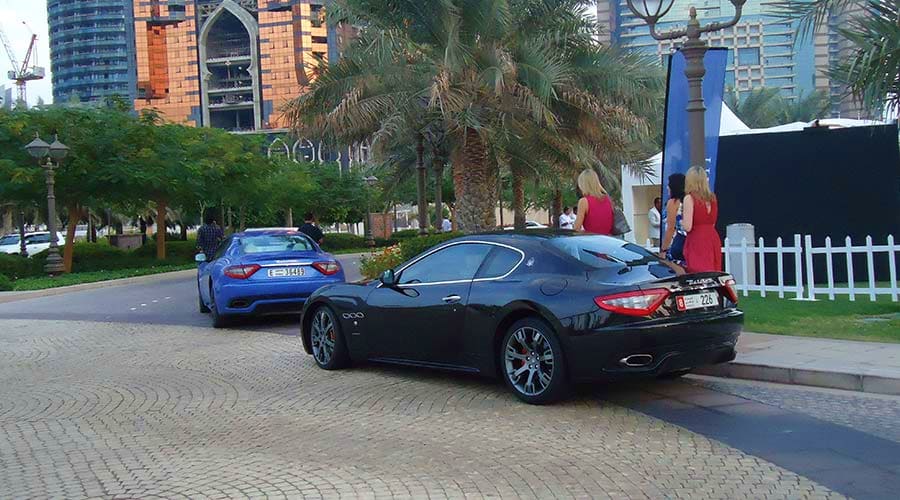Abu Dhabi to Dubai Intercity transfers Black Maserati and blue sports car parked on palm-lined boulevard with modern high-rise buildings and waterfront promenade in
