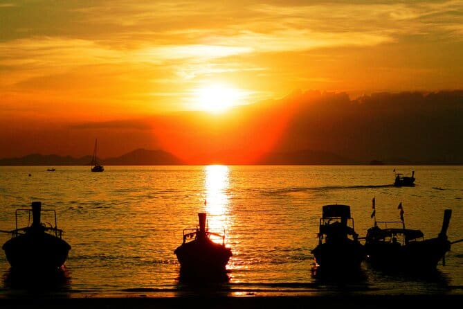 Golden sunset over calm water with silhouetted long tail boats anchored near shore and mountainous island backdrop