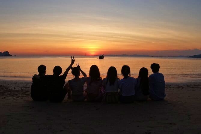 Group of tourists sitting on beach at sunset, facing calm waters with longtail boat visible on horizon