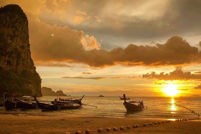 Traditional long tail boats moored on beach at sunset with limestone cliff and golden sky reflecting on water