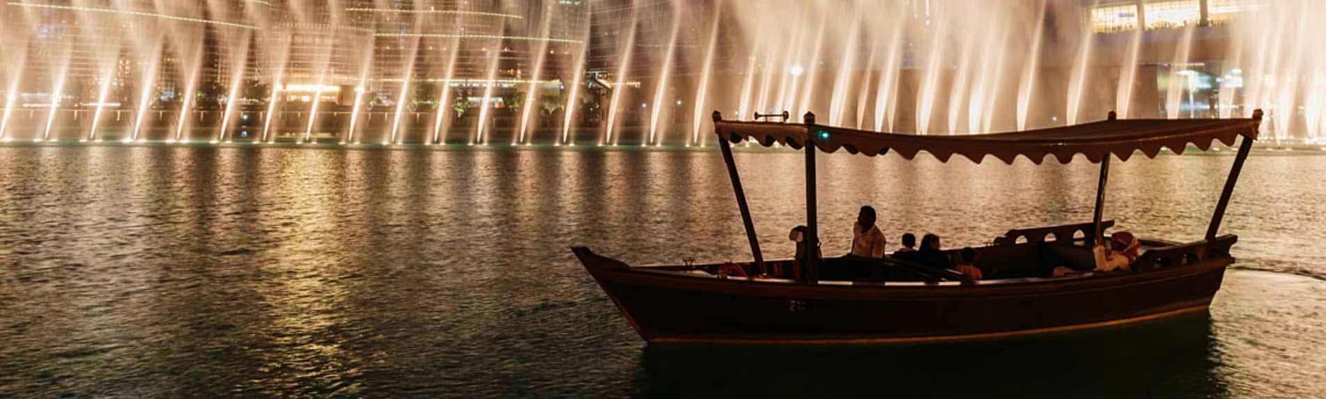 Dubai Fountain Lake Ride from Abu Dhabi Traditional wooden abra boat with passengers on Dubai lake at night with illuminated fountain and modern building in backgrou