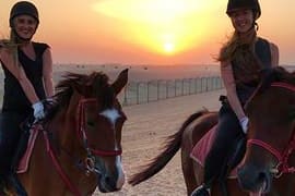 Two women riding horses on a beach at sunset with golden sky and ocean in background