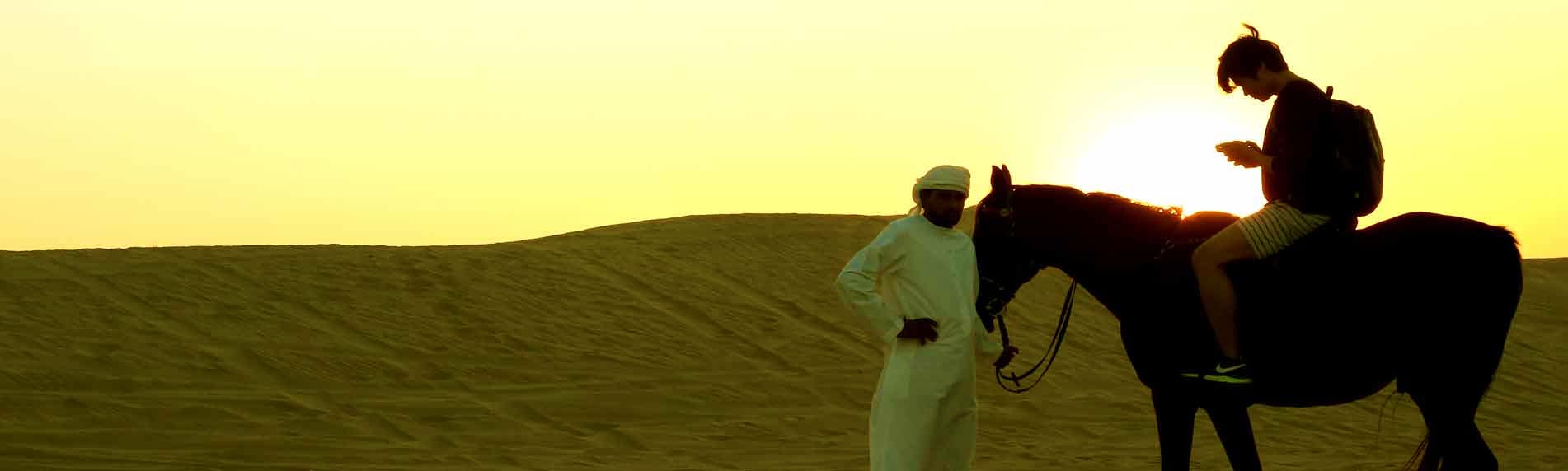 Child riding a brown horse in desert dunes at sunset with guide holding reins