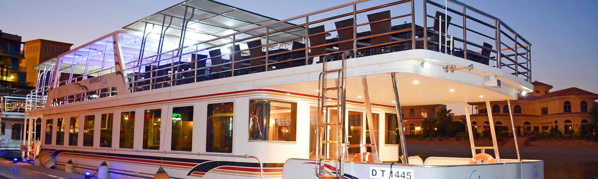 Modern two-deck white cruise ship illuminated at dusk with open-air upper deck and large windows, docked at waterfront promen