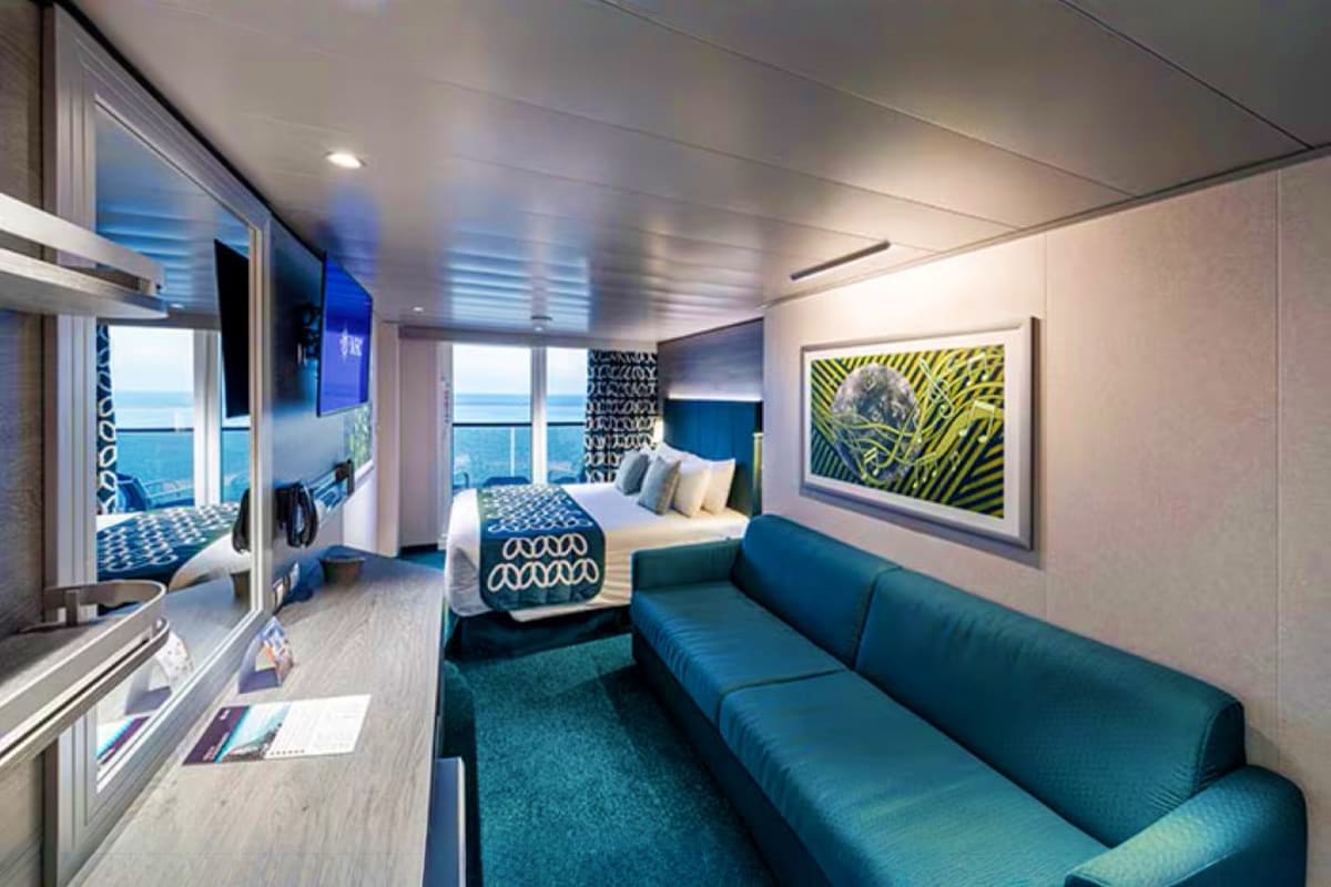 Modern cruise ship cabin with blue sofa, queen bed, ocean view windows, and contemporary furnishings