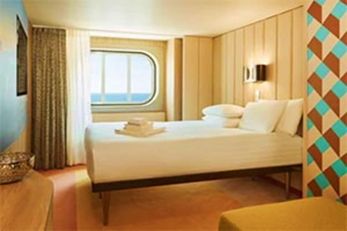 Modern cruise ship cabin with twin bed, porthole window, cream and tan décor, and geometric patterned accent wall