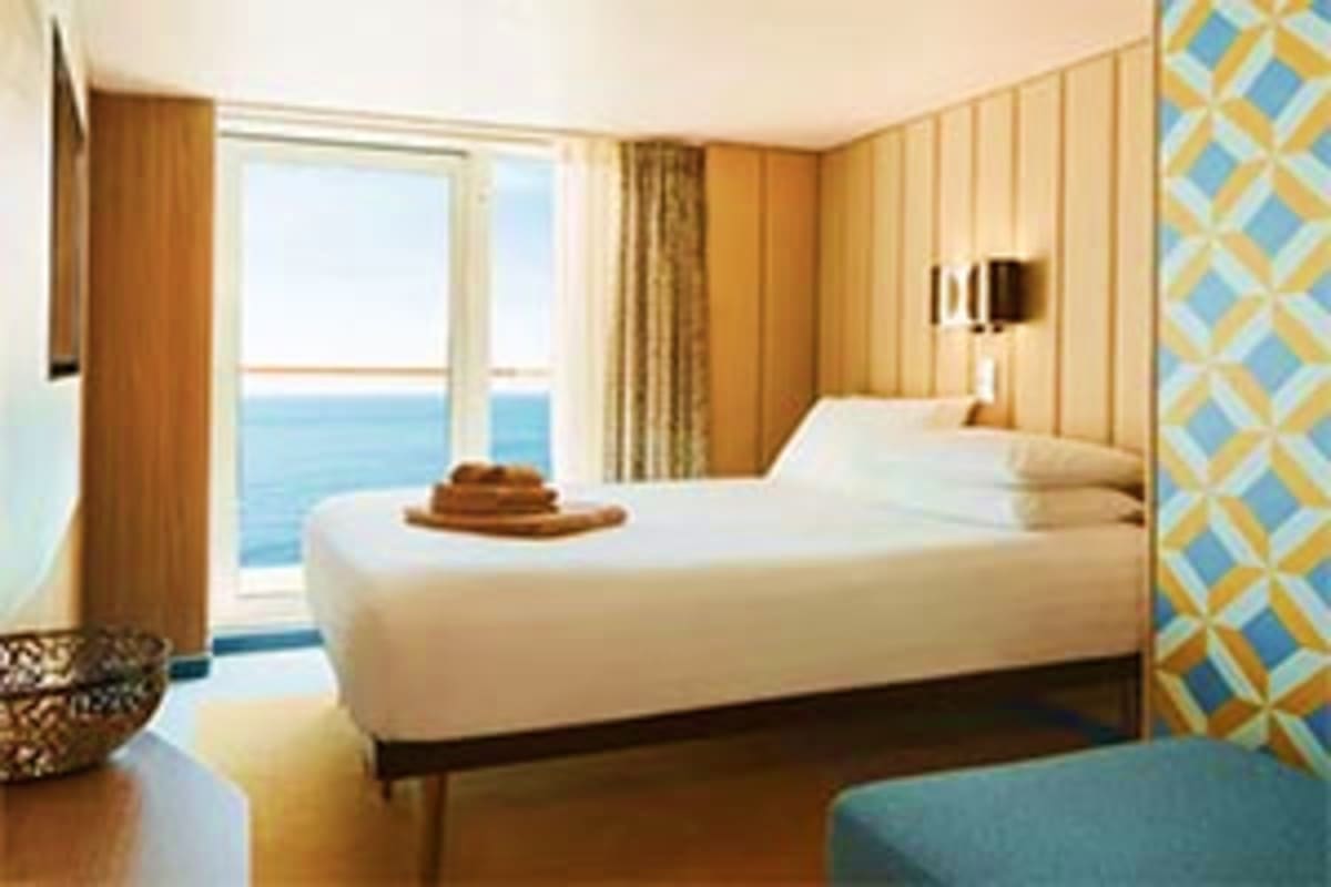 Luxury cruise ship cabin with ocean view balcony, white bed, warm wood paneling, and modern furnishings overlooking blue Medi