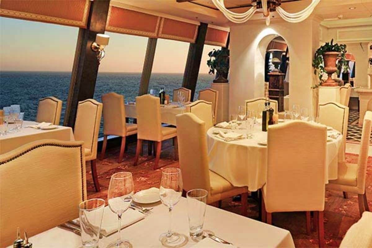 Elegant ship dining room with round tables, beige chairs, and panoramic ocean views through large windows at sunset
