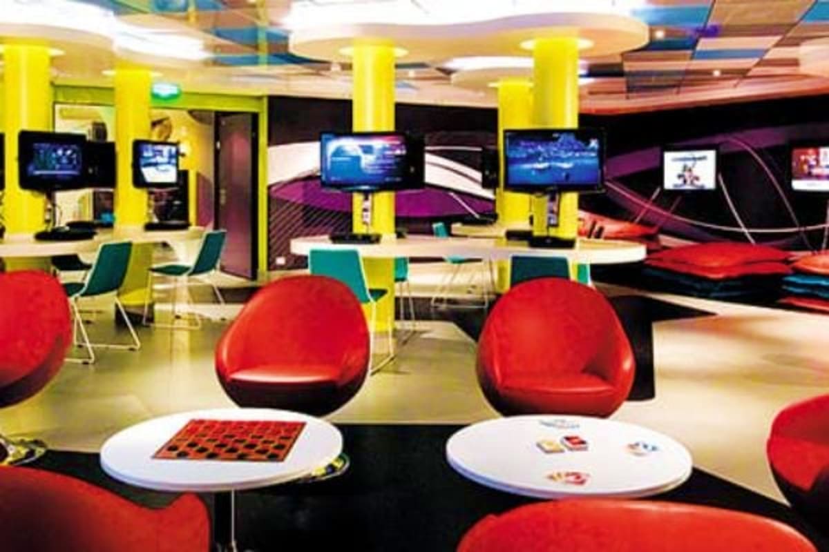 Modern cruise ship entertainment lounge with red pod-style seating, white tables with board games, computer workstations, and