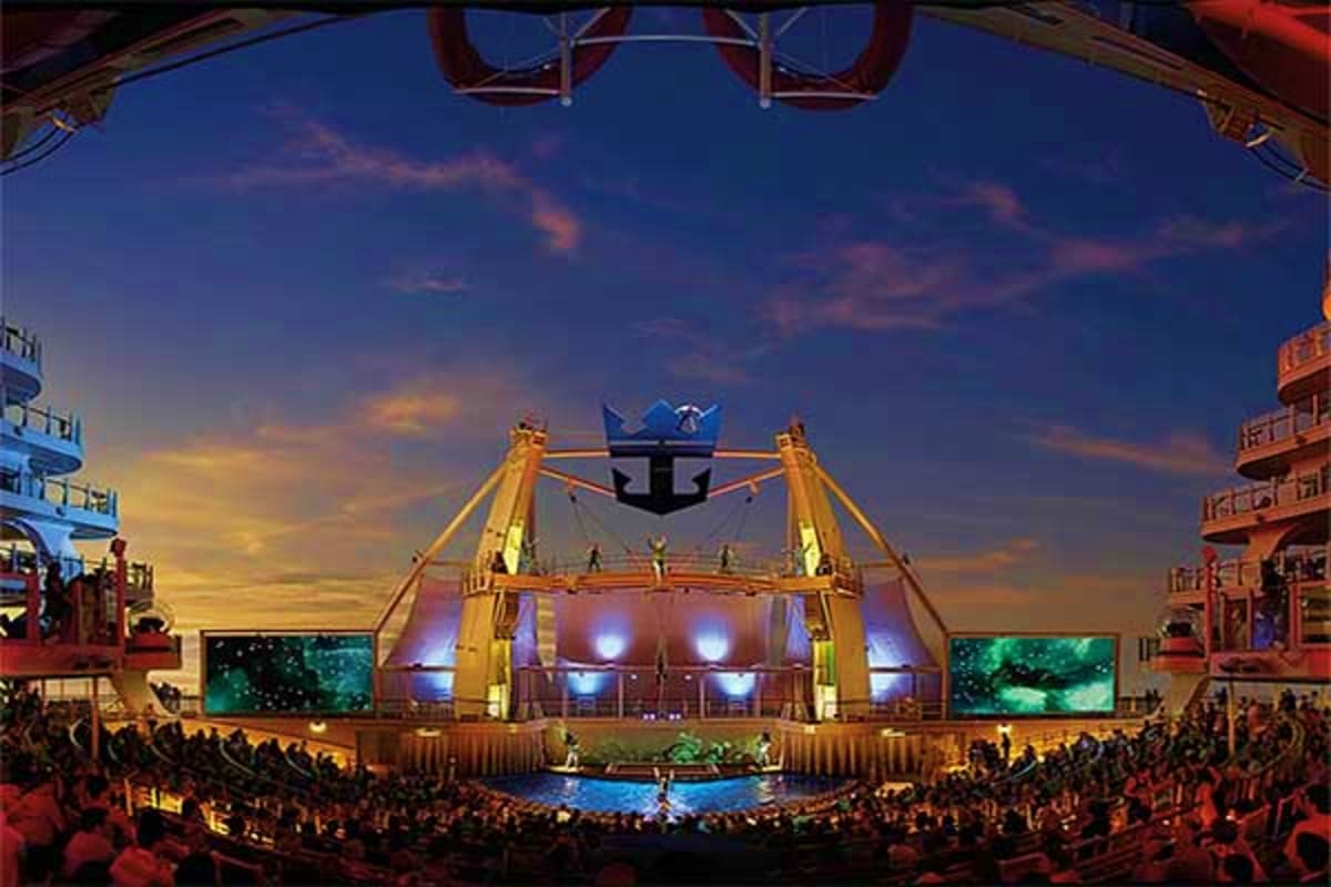 Large illuminated stage with blue crown logo suspended above, surrounded by packed audience on Royal Caribbean cruise ship at