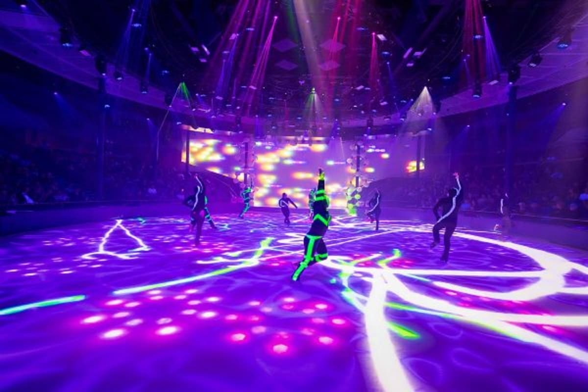 Performers in neon green outfits dancing on illuminated purple floor stage with geometric light patterns and hanging architec