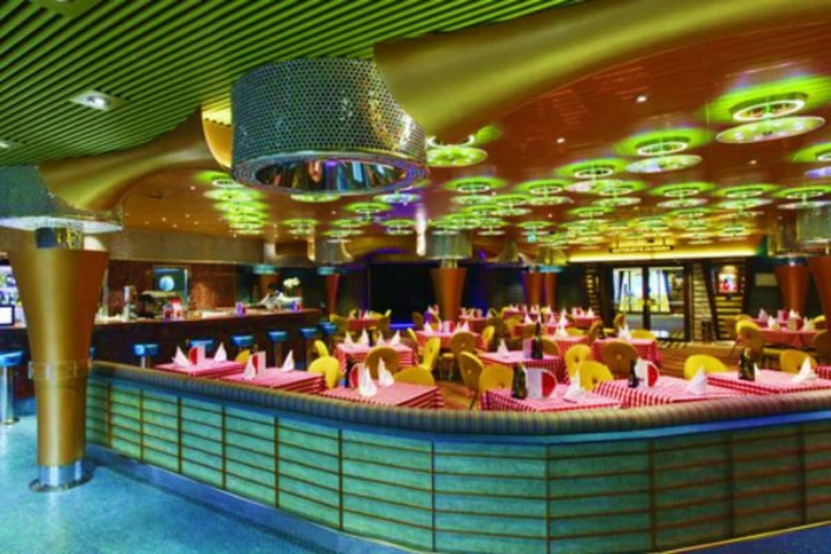 Modern cruise ship dining room with yellow chairs, pink tablecloths, green ceiling lights, and service counter
