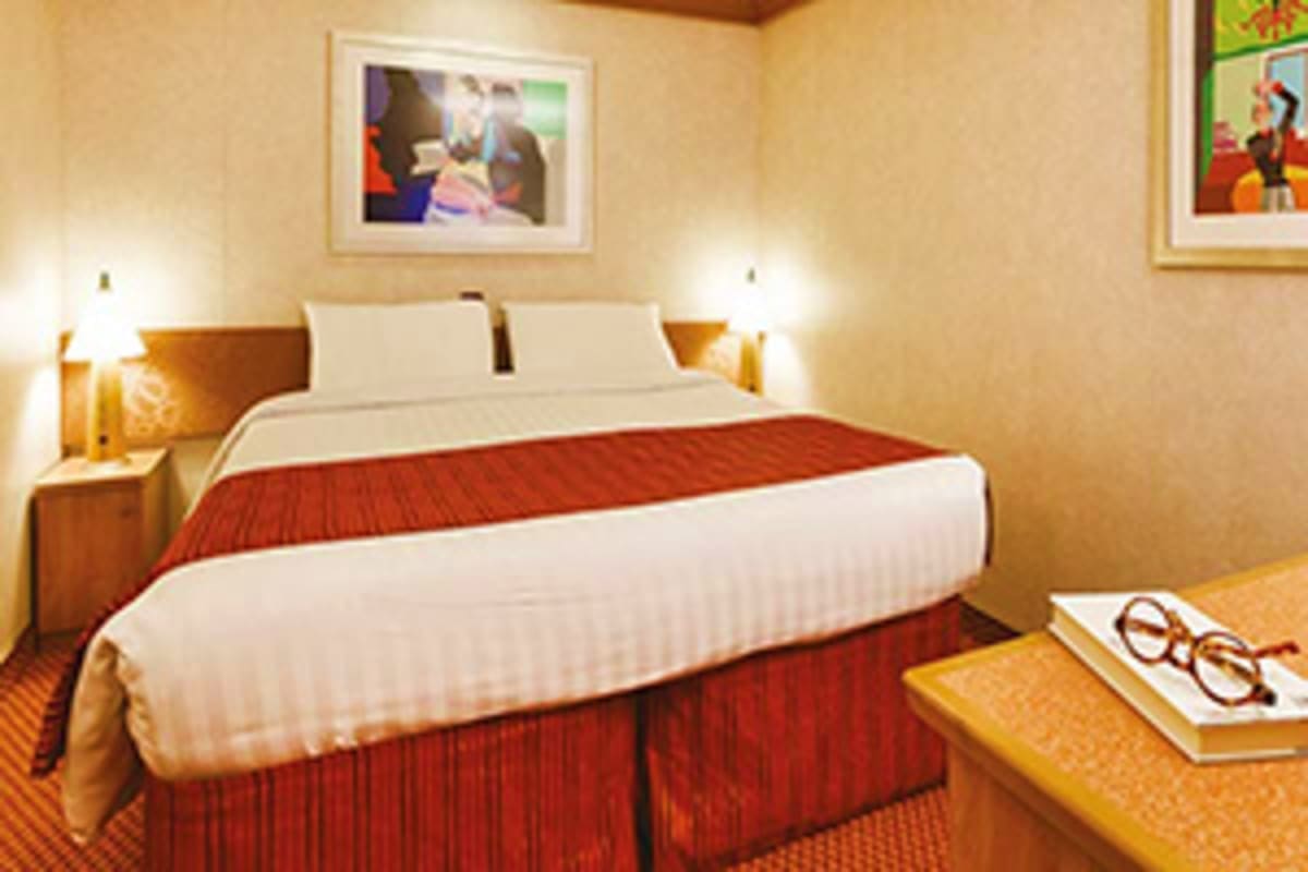 Modern hotel bedroom with red and white bedding, wooden nightstands with lamps, framed artwork on beige walls, and orange car