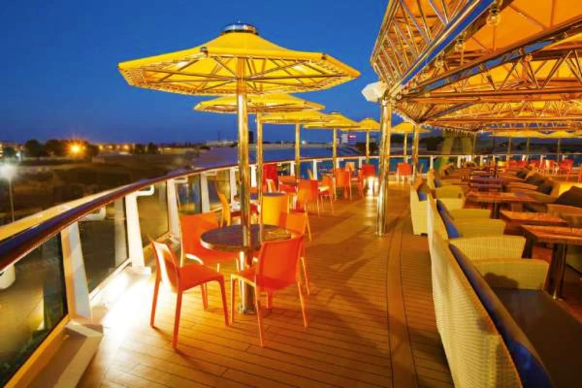 Illuminated outdoor deck of cruise ship at dusk with yellow umbrellas, orange chairs, and river view at twilight