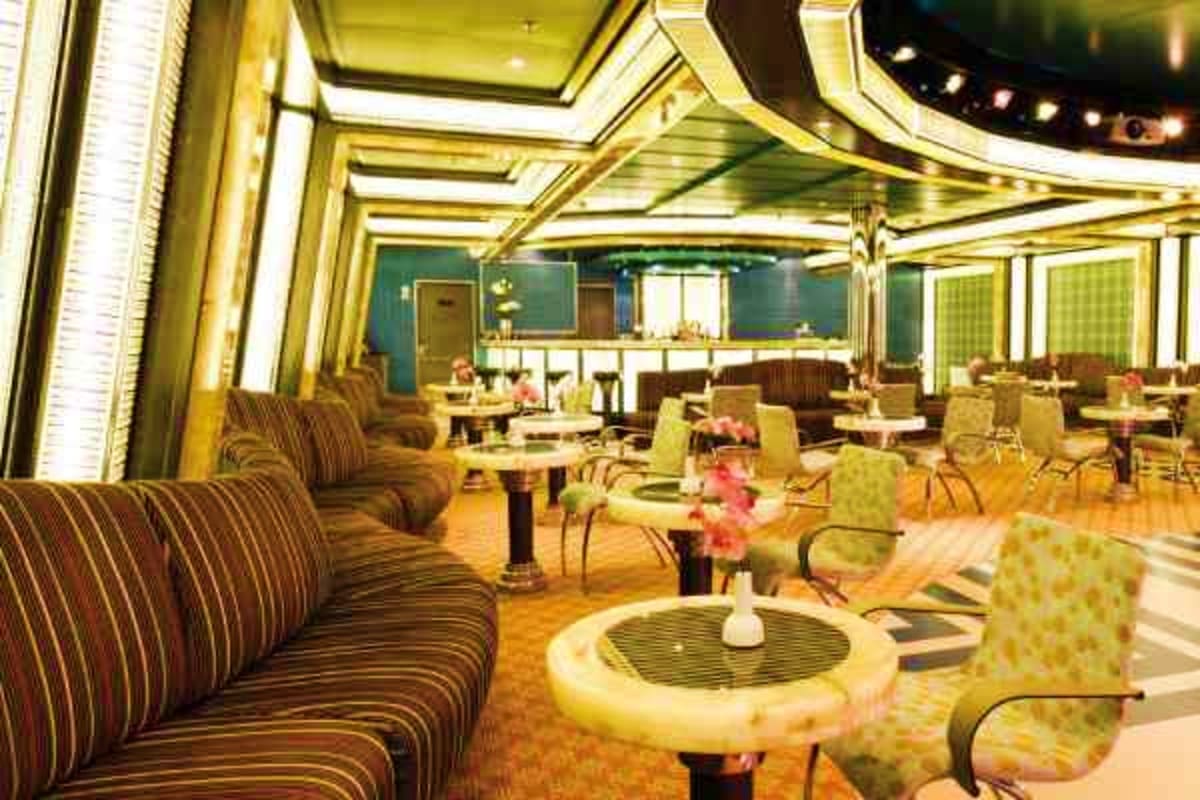 Elegant ship dining room with striped upholstered seating, round tables with pink floral centerpieces, gold ceiling details, 