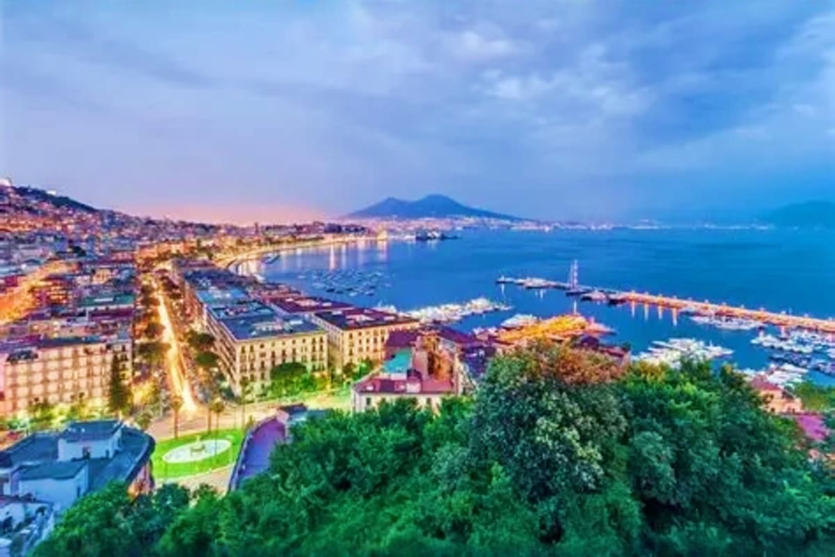 Aerial view of Naples coastline at dusk with Mount Vesuvius in the distance, illuminated buildings, harbor with boats, and bl