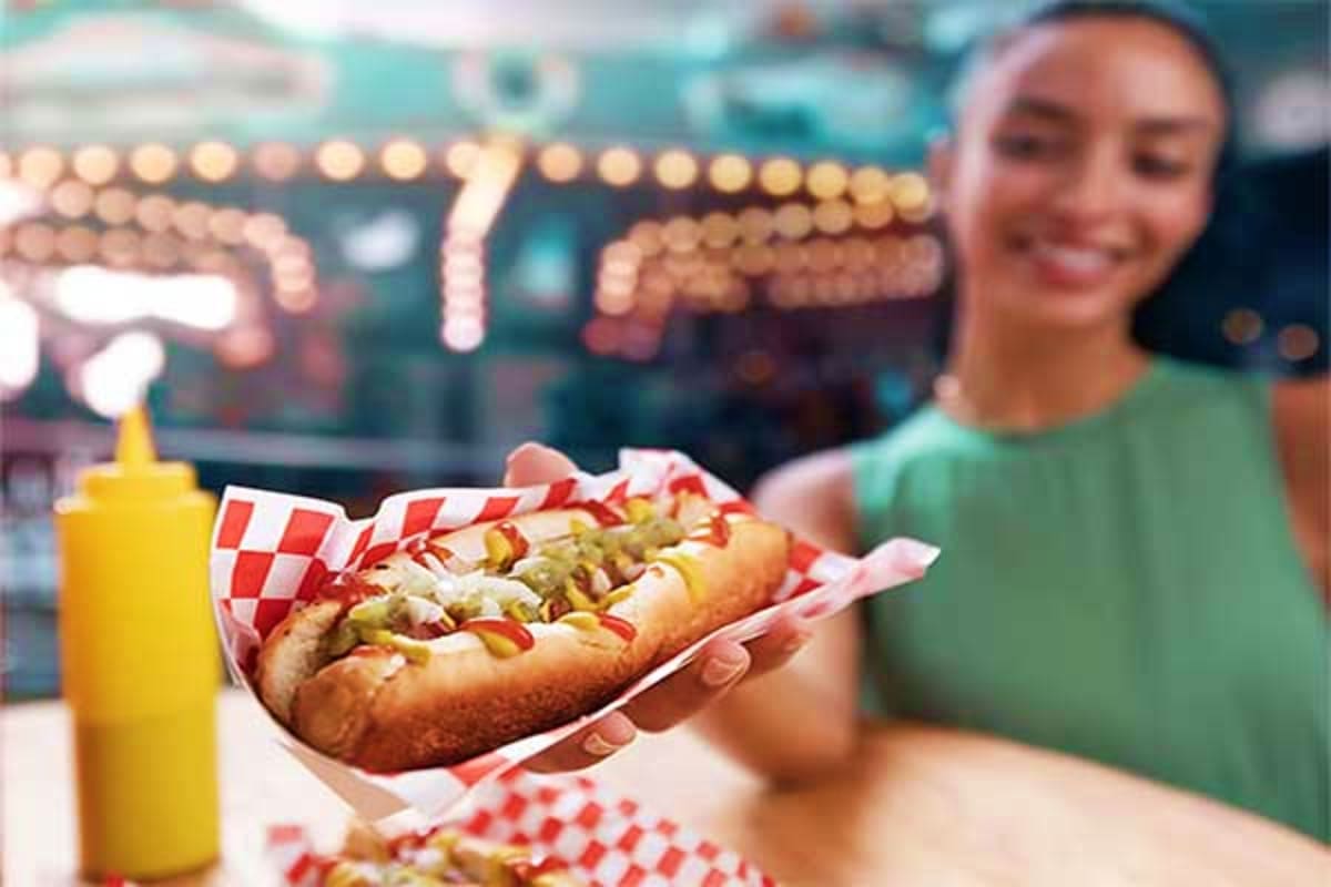 Woman holding hot dog with mustard and relish at outdoor carnival stand with illuminated signage in background