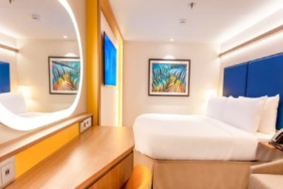Modern cruise ship cabin with blue headboard bed, illuminated mirror, yellow accent trim, and tropical artwork on walls