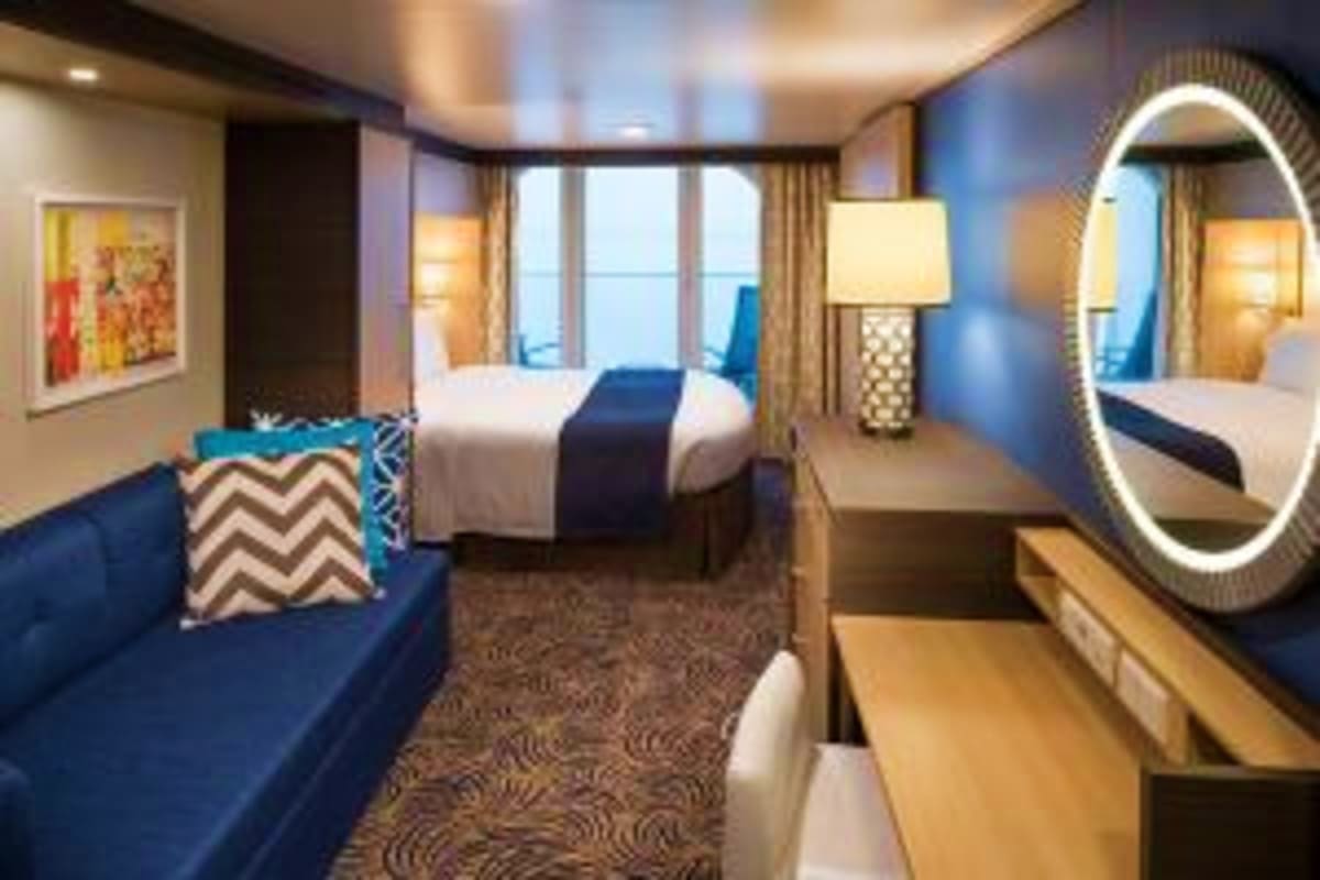 Modern cruise ship stateroom with queen bed, blue sofa, illuminated oval mirror, and ocean view windows