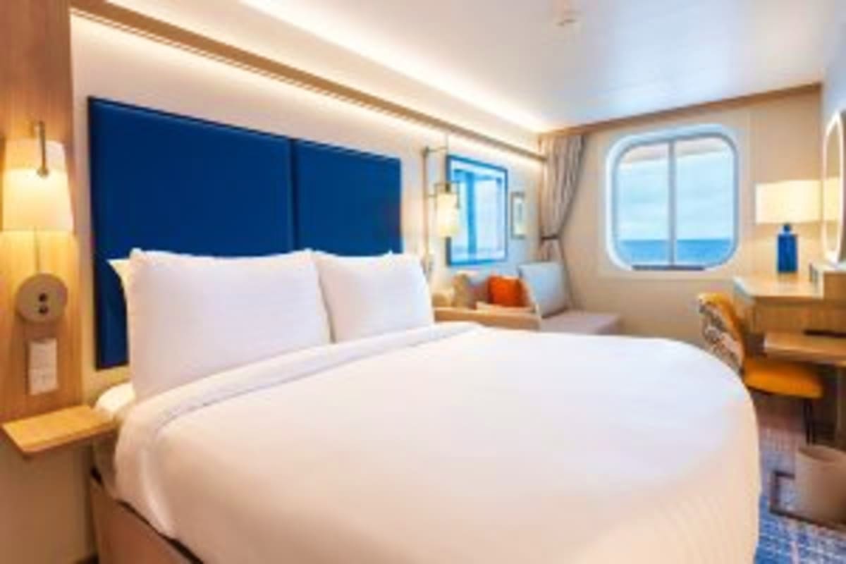 Modern cruise ship cabin with queen bed, blue upholstered headboard, porthole window overlooking ocean, and contemporary furn