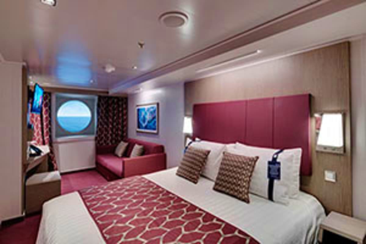 Luxury cruise ship cabin with queen bed, burgundy headboard, porthole window, seating area, and modern décor