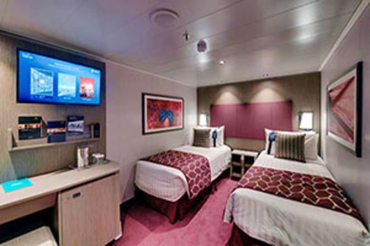 Modern cruise ship interior stateroom with two beds, burgundy accents, flat-screen TV, desk area, and porthole windows