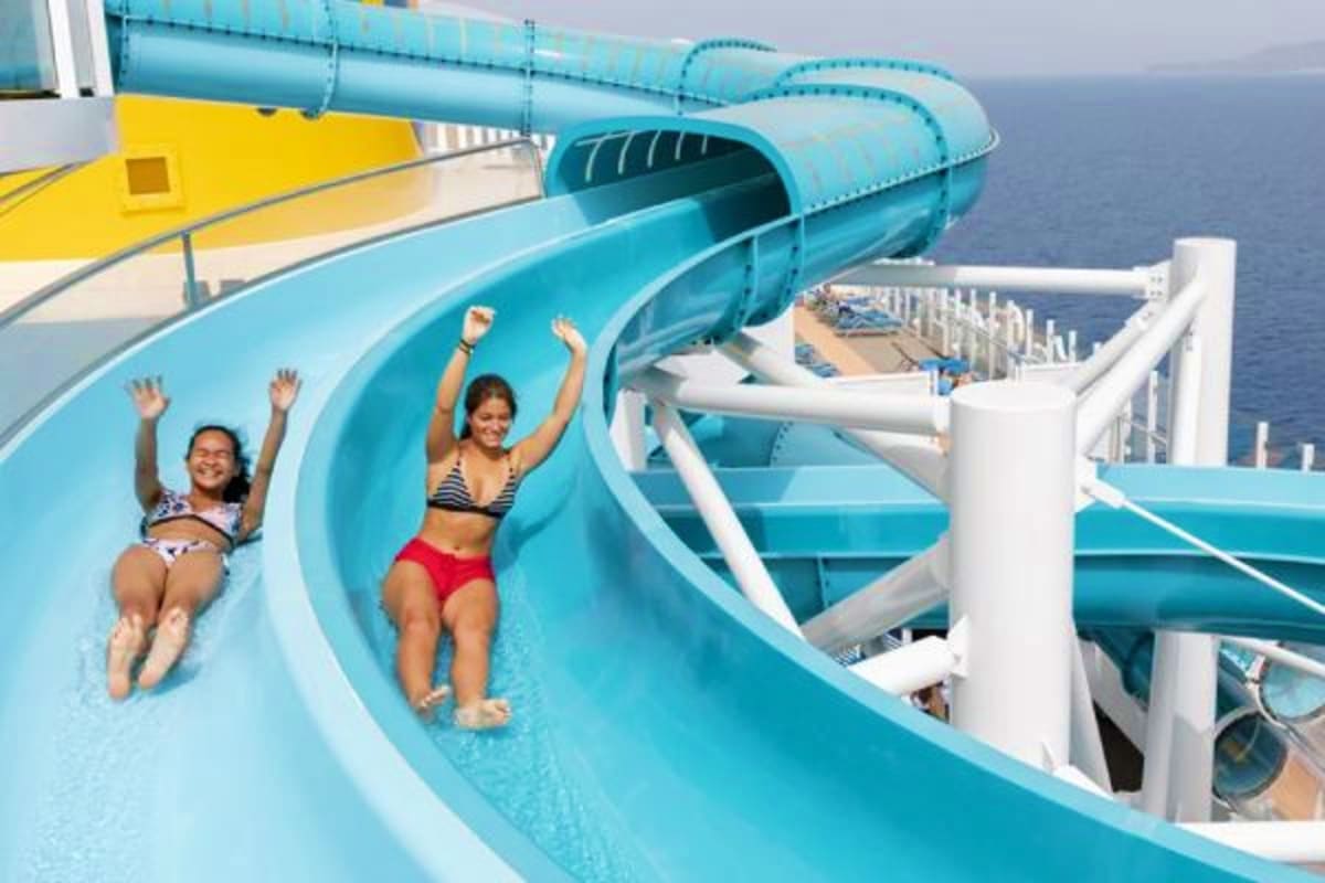 Two children sliding down a bright turquoise water slide on a cruise ship with ocean visible in background