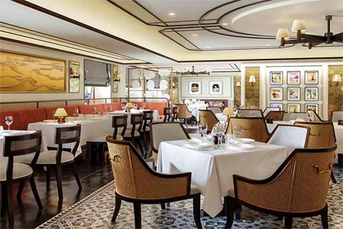 Elegant cruise ship dining room with white tablecloth-covered tables, woven chairs, red banquette seating, and framed artwork