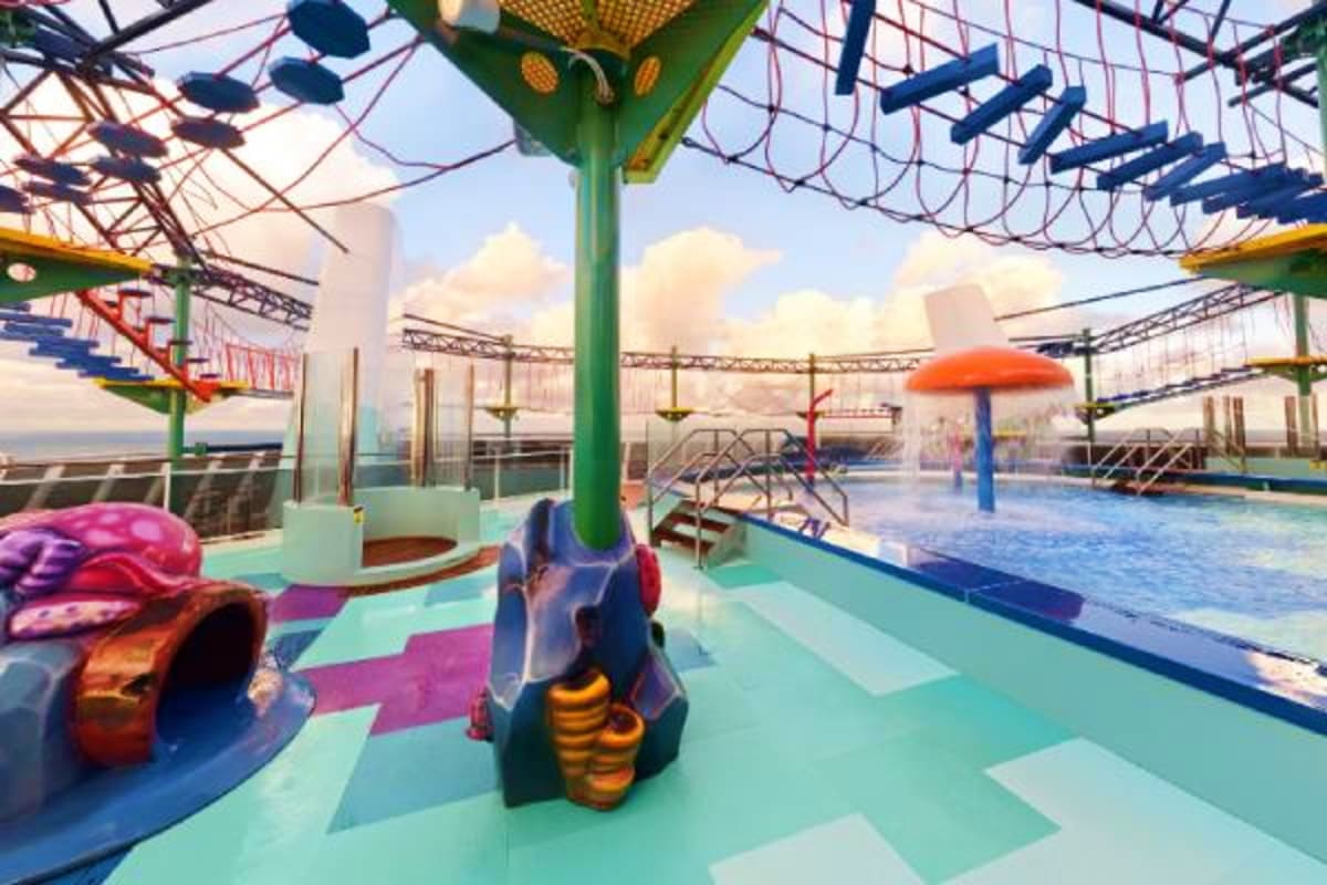 Colorful kids water park area on cruise ship deck with climbing structures, pool, water slides, and ocean cityscape in backgr